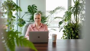 Tine Løvik sitter i et lyst, frodig rom fylt med planter med laptop og kaffe.
